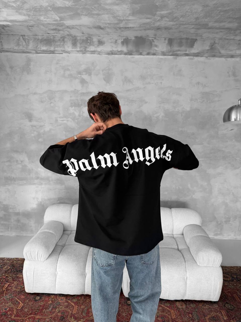 A person stands facing away, wearing a Palm Angels Style Cotton T-Shirt featuring a bold white gothic back print. They are in front of a light gray couch and textured wall, with a patterned rug beneath them.