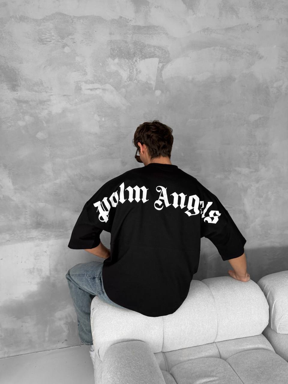 A person with short brown hair, wearing a black Palm Angels Style Cotton T-Shirt featuring a gothic back print and light blue jeans, sits on the arm of a white sofa facing a gray textured wall.