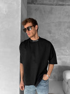 A man in dark sunglasses wears a Palm Angels Style Cotton T-Shirt with a black back print, paired with loose black and light blue jeans, leaning casually against a minimalist gray concrete wall with one hand in his pocket.