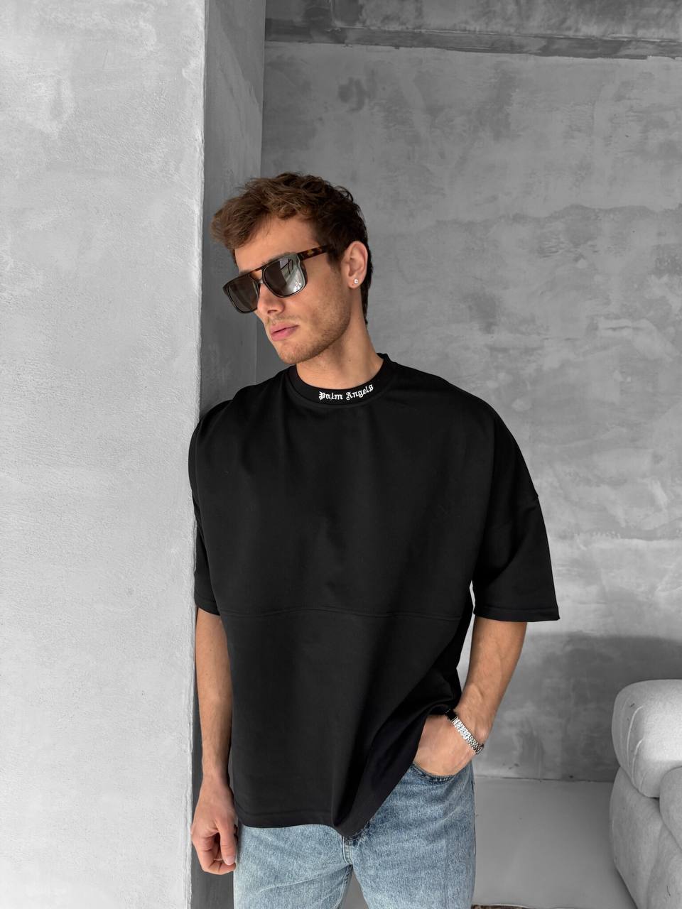 A man in dark sunglasses wears a Palm Angels Style Cotton T-Shirt with a black back print, paired with loose black and light blue jeans, leaning casually against a minimalist gray concrete wall with one hand in his pocket.
