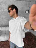 A young man in sunglasses and a Palm Angels Style Cotton T-Shirt takes a selfie, standing in a modern room with a white sofa, gray textured wall, floor lamp, and red patterned rug.