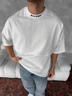 A man stands indoors wearing a loose white Palm Angels Style Cotton T-Shirt with black collar lettering and a gothic back print, paired with light blue jeans and a watch. A couch and red-patterned rug are in the background.