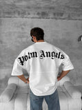 A person with their back to the camera wears a Palm Angels Style Cotton T-Shirt featuring a bold gothic print, standing in a modern gray-toned room with a white sofa.