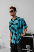 A man wearing the Tommy Vercetti Oversize Tropical Shirt – Palm Print Button-Up and black sunglasses stands indoors with one hand in his pocket, near a black equipment case and a metal chair.