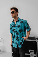 A man wearing the Tommy Vercetti Oversize Tropical Shirt – Palm Print Button-Up and black sunglasses stands indoors with one hand in his pocket, near a black equipment case and a metal chair.