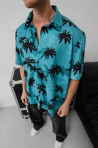 A man wears the Tommy Vercetti Oversize Tropical Shirt – Palm Print Button-Up, standing indoors next to a black couch and metal case. His blue shirt features a bold black palm tree print, and his face is partially out of frame.