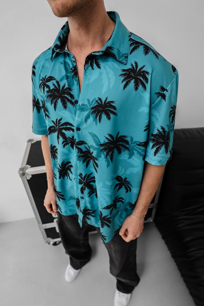 A man wears the Tommy Vercetti Oversize Tropical Shirt – Palm Print Button-Up, standing indoors next to a black couch and metal case. His blue shirt features a bold black palm tree print, and his face is partially out of frame.