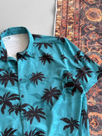 A Tommy Vercetti Oversize Tropical Shirt – Palm Print Button-Up in teal lies by the corner of a multicolored patterned rug on a light gray surface.
