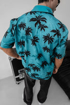 Wearing the Tommy Vercetti Oversize Tropical Shirt – Palm Print Button-Up, a person with dark hair stands with their back to the camera near a black case and sofa in a bright room.