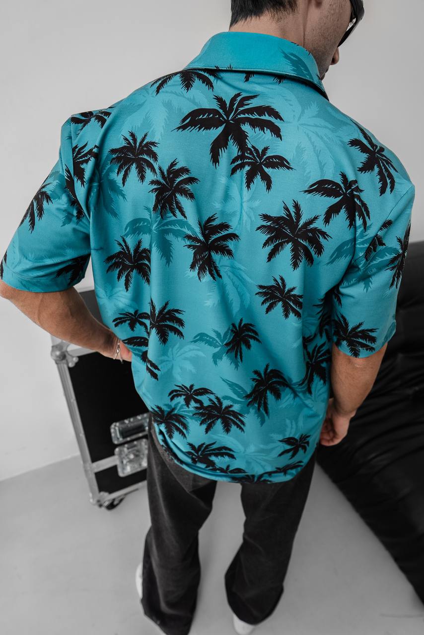 Wearing the Tommy Vercetti Oversize Tropical Shirt – Palm Print Button-Up, a person with dark hair stands with their back to the camera near a black case and sofa in a bright room.