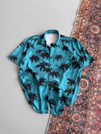 The Tommy Vercetti Oversize Tropical Shirt – Palm Print Button-Up, in blue with black palm tree print, is laid flat across a patterned rug and smooth gray surface for a relaxed, stylish vibe.