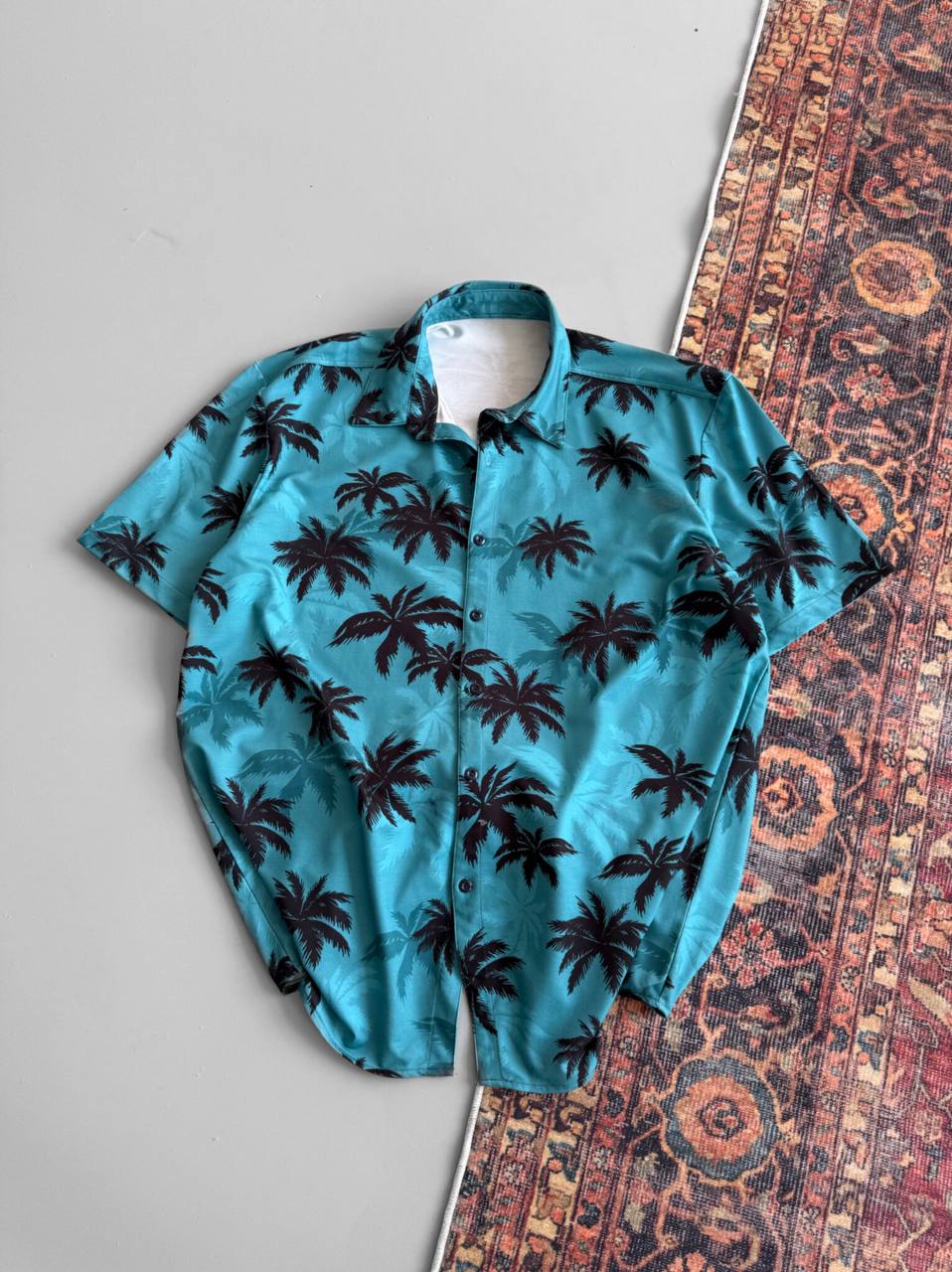 The Tommy Vercetti Oversize Tropical Shirt – Palm Print Button-Up, in blue with black palm tree print, is laid flat across a patterned rug and smooth gray surface for a relaxed, stylish vibe.