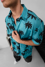 A man stands indoors wearing the Tommy Vercetti Oversize Tropical Shirt – Palm Print Button-Up in turquoise. He holds the collar with one hand, his face partly visible, against a minimal, bright background.