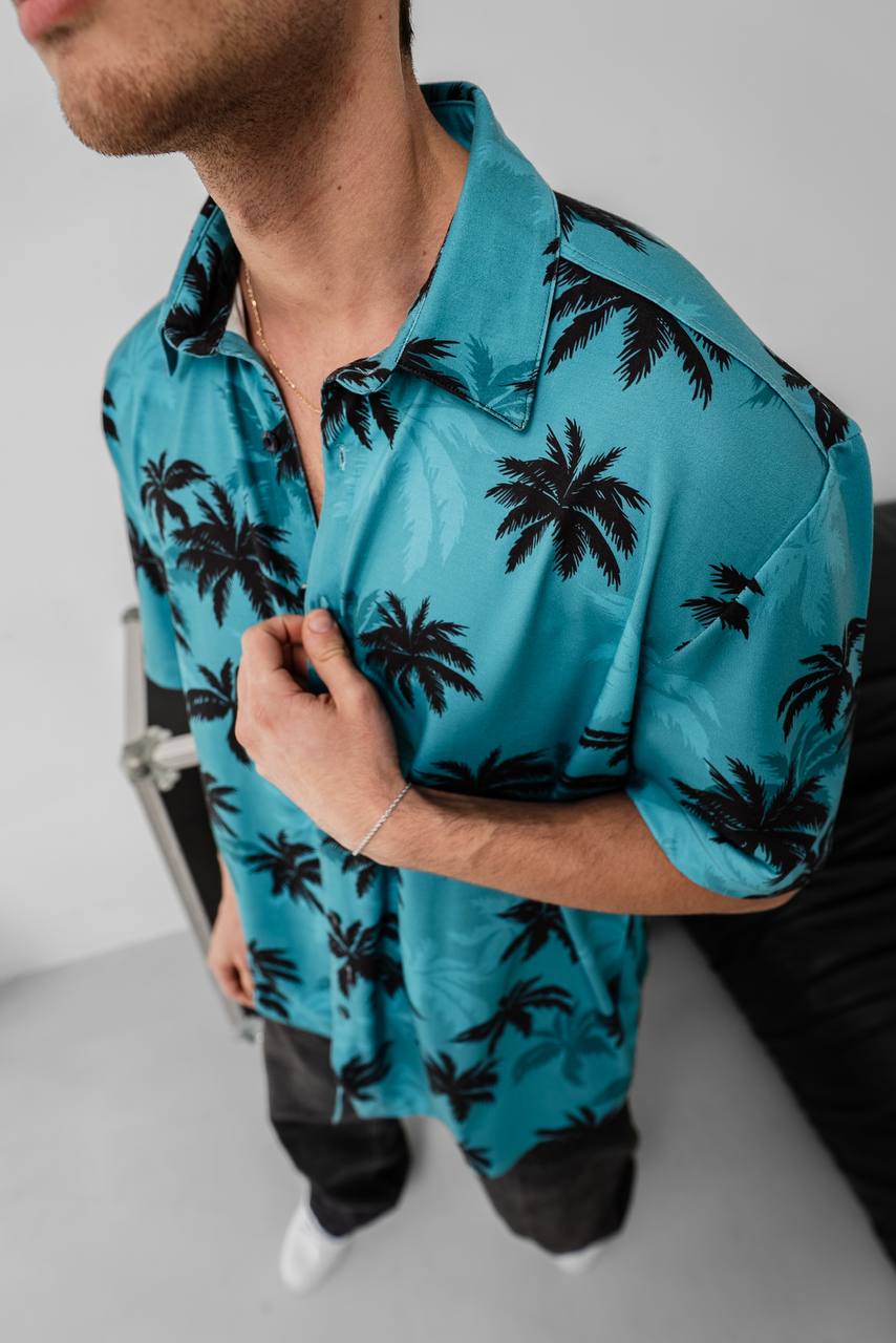 A man stands indoors wearing the Tommy Vercetti Oversize Tropical Shirt – Palm Print Button-Up in turquoise. He holds the collar with one hand, his face partly visible, against a minimal, bright background.