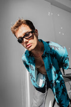 A young man in black sunglasses and a Tommy Vercetti Oversize Tropical Shirt – Palm Print Button-Up poses confidently indoors, leaning forward as sunlight casts shadows on the white wall behind him.