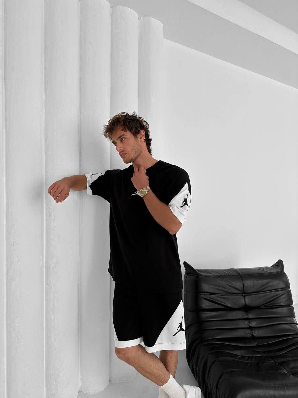 A man leans against a white column in a minimalist room, wearing the Black & White Jordan-Inspired Two Piece Set – Men’s Casual Sportswear Outfit with white socks. A black leather couch beside him adds to the laid-back vibe.