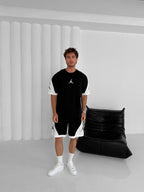 A man wears the Black & White Jordan-Inspired Two Piece Set – Men’s Casual Sportswear Outfit, featuring a T-shirt and shorts. He stands by a black leather chair in a minimalistic white room with vertical wall paneling.