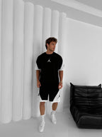 A man stands indoors beside a black leather chair, wearing the Black & White Jordan-Inspired Two Piece Set – Men’s Casual Sportswear Outfit with white sneakers, creating a modern streetwear look against a white paneled wall.