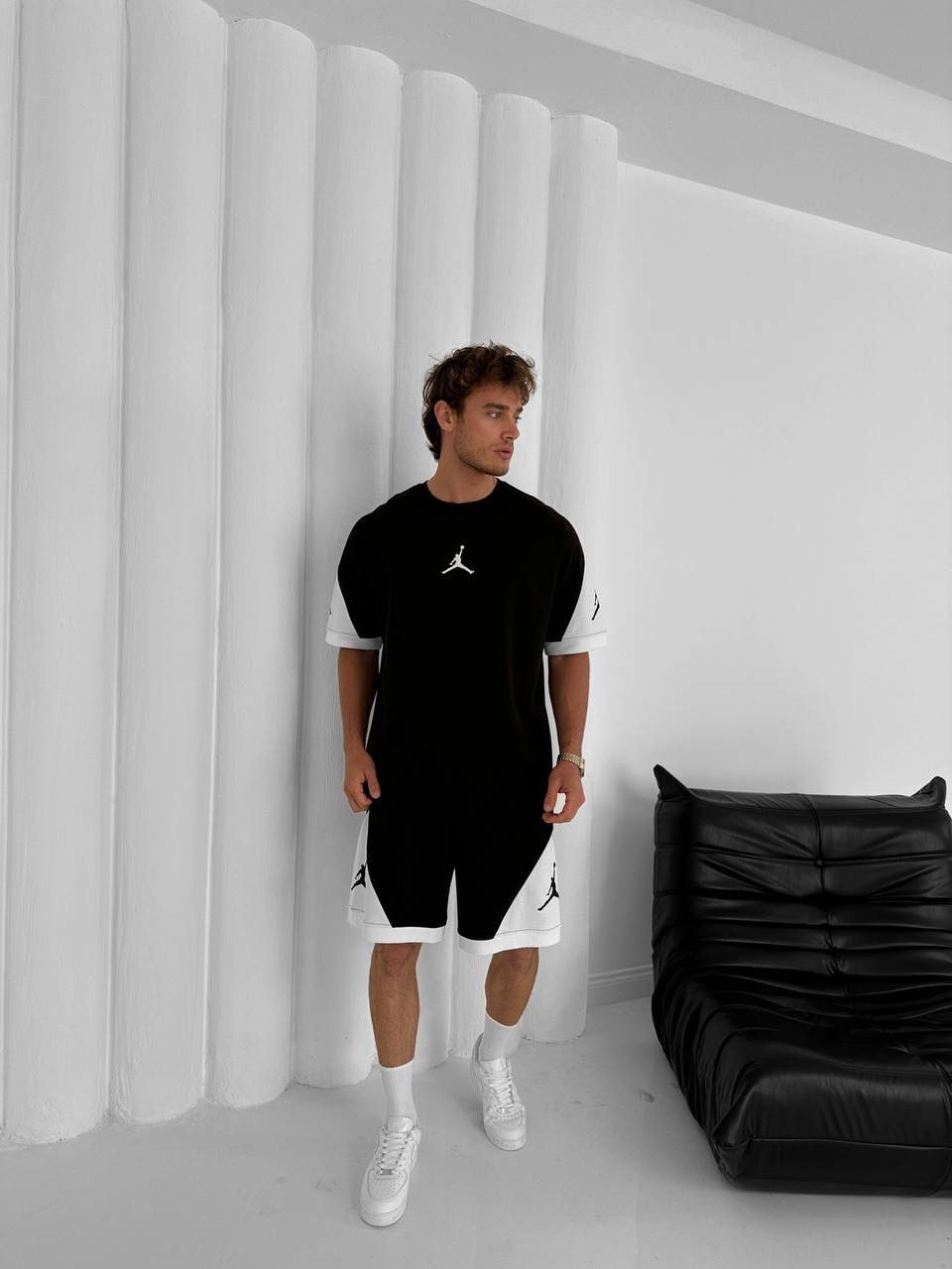 A man stands indoors beside a black leather chair, wearing the Black & White Jordan-Inspired Two Piece Set – Men’s Casual Sportswear Outfit with white sneakers, creating a modern streetwear look against a white paneled wall.