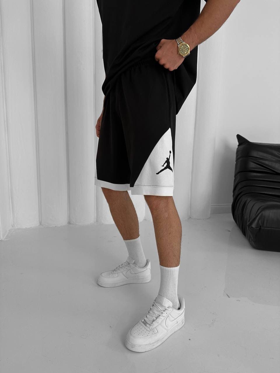 Wearing the Black & White Jordan-Inspired Two Piece Set – Men’s Casual Sportswear Outfit, a person stands indoors by a black sofa. Only the lower half of their streetwear look is visible: shorts with the Jordan logo, white socks, and sneakers.