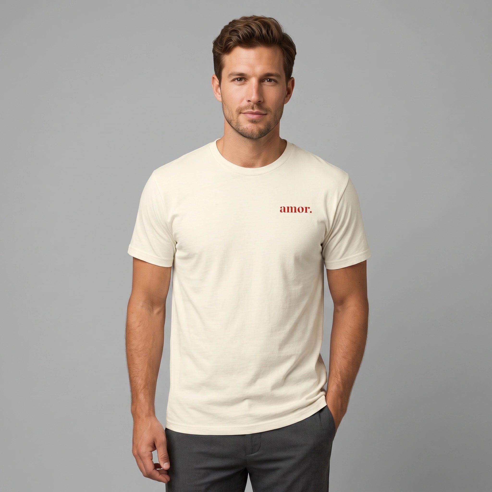 A front-view photograph of a male model, from the waist up, wearing a short-sleeved, crew-neck t-shirt in a faded cream color. The t-shirt features a small, single word in red text, "amor.", printed on the left chest. The model is standing, facing forward, with his left hand in his trouser pocket, and has a direct, calm expression. He is wearing charcoal grey trousers. The background is a plain, neutral light grey.