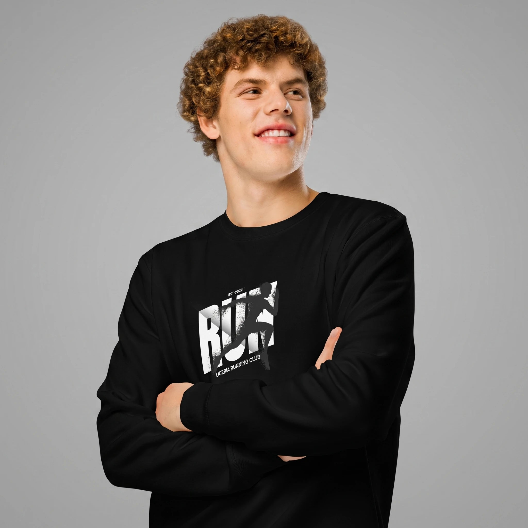 Close-up front view of a smiling male model with curly brown hair posing with crossed arms against a solid grey background. He is wearing a black crewneck sweatshirt featuring a chest print with the bold text 'RUN', a runner's silhouette over the typography, and 'LICERIA RUNNING CLUB' printed beneath.