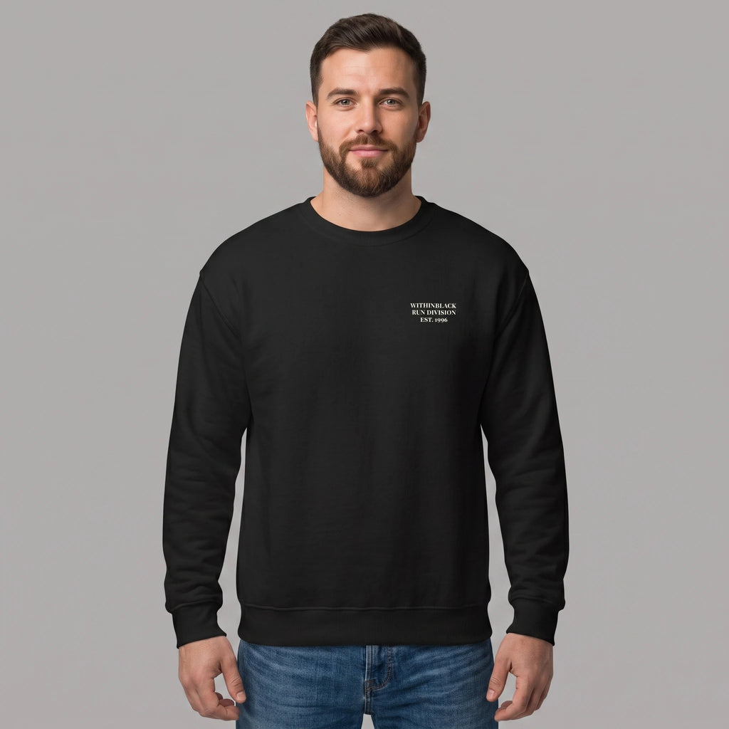Front view of a smiling bearded male model standing against a grey studio background, wearing a black crewneck sweatshirt and blue jeans. The sweatshirt features small white text printed on the left chest reading 'WITHINBLACK RUN DIVISION EST. 1996'.