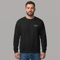 Front view of a smiling bearded male model standing against a grey studio background, wearing a black crewneck sweatshirt and blue jeans. The sweatshirt features small white text printed on the left chest reading 'WITHINBLACK RUN DIVISION EST. 1996'.