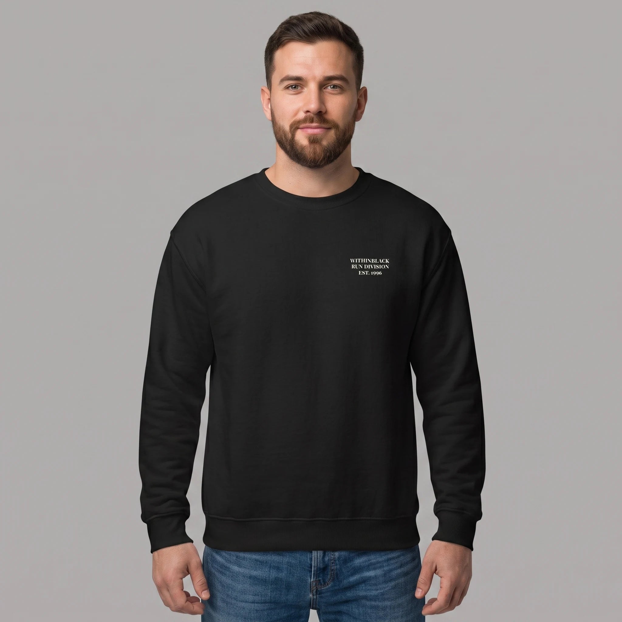 Front view of a smiling bearded male model standing against a grey studio background, wearing a black crewneck sweatshirt and blue jeans. The sweatshirt features small white text printed on the left chest reading 'WITHINBLACK RUN DIVISION EST. 1996'.