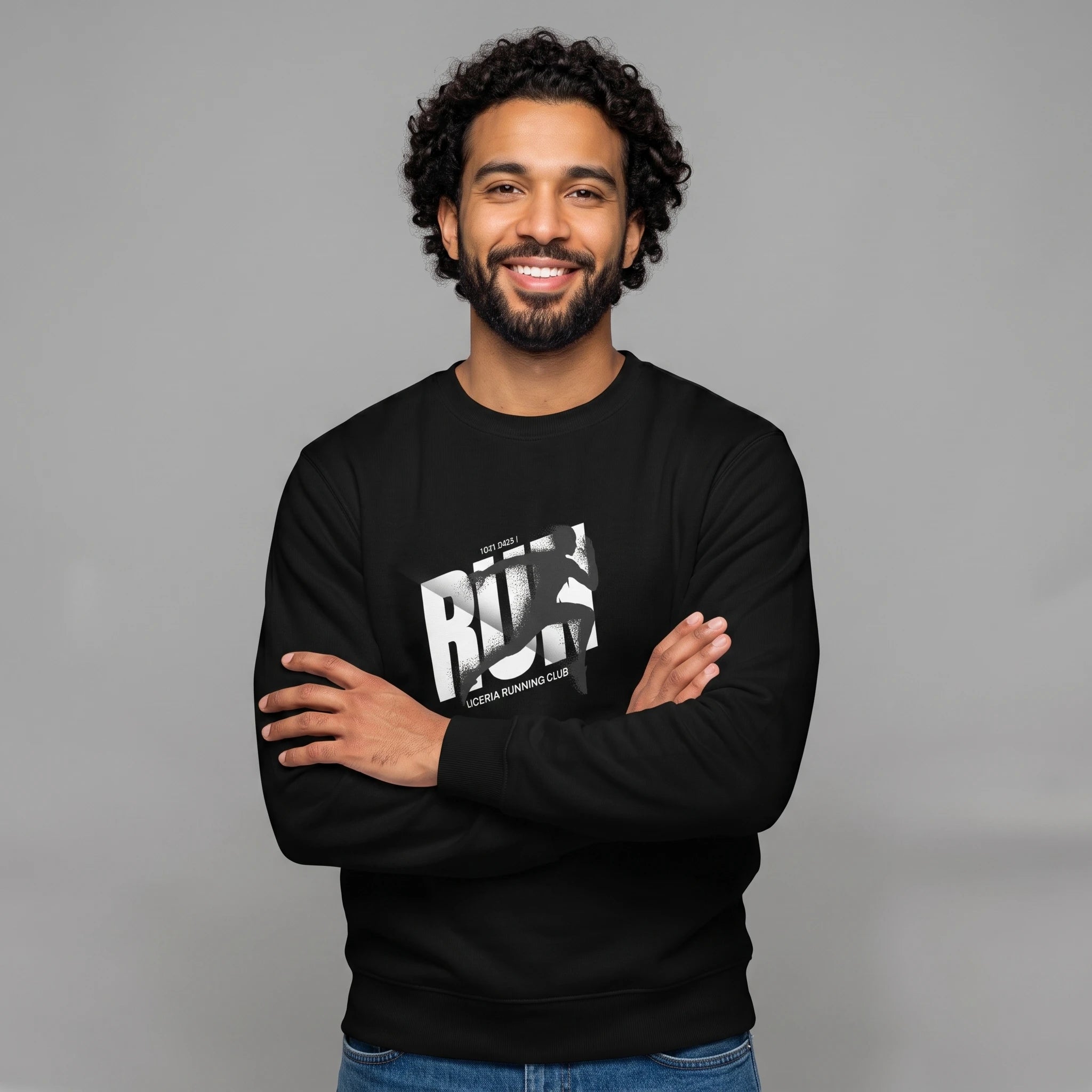 Front-facing studio shot of a smiling male model with dark curly hair and a beard, standing with arms crossed against a grey background. He is wearing a black crewneck sweatshirt with blue jeans, featuring a chest graphic of bold white 'RUN' lettering, a sprinting runner silhouette overlay, and 'LICERIA RUNNING CLUB' text printed below.