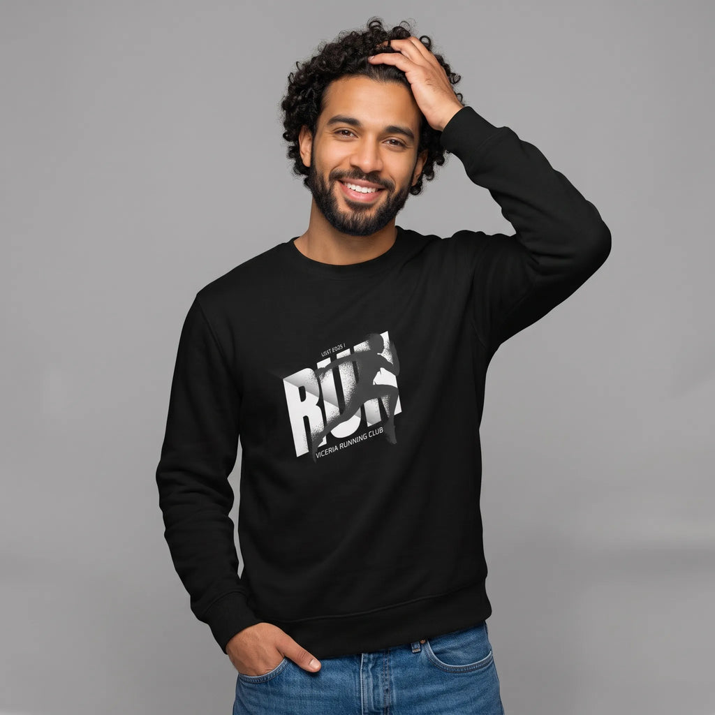 Smiling male model with dark curly hair and a beard, standing against a grey background with one hand touching his head and the other in his blue jeans pocket. He is wearing a black crewneck sweatshirt featuring a chest print with bold white 'RUN' letters, a dark runner's silhouette over the text, and 'LICERIA RUNNING CLUB' printed below.