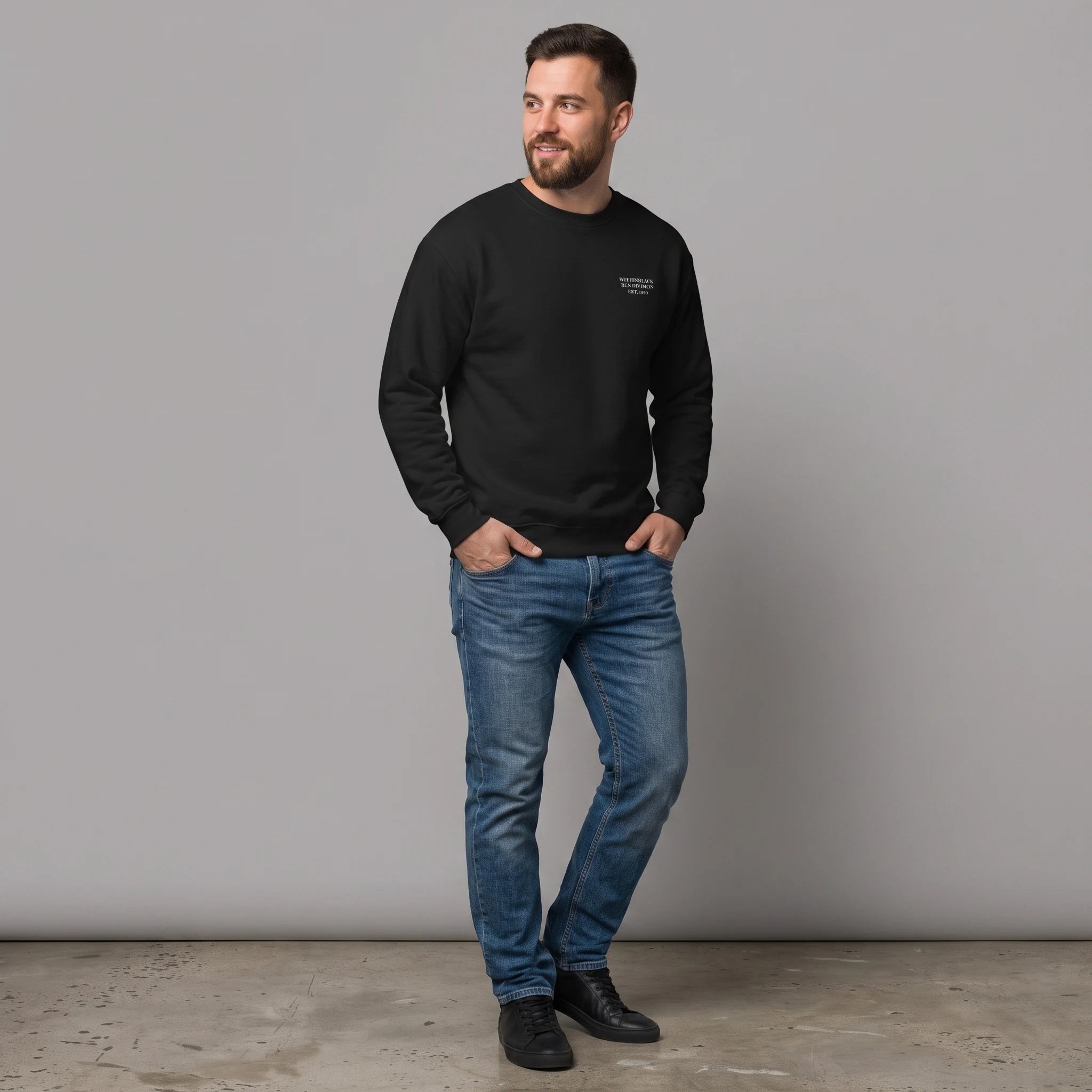 Full-length front view of a smiling bearded male model standing against a grey studio background with his hands in his pockets. He is wearing a black crewneck sweatshirt with small white text printed on the left chest, paired with blue slim-fit jeans and black leather sneakers.