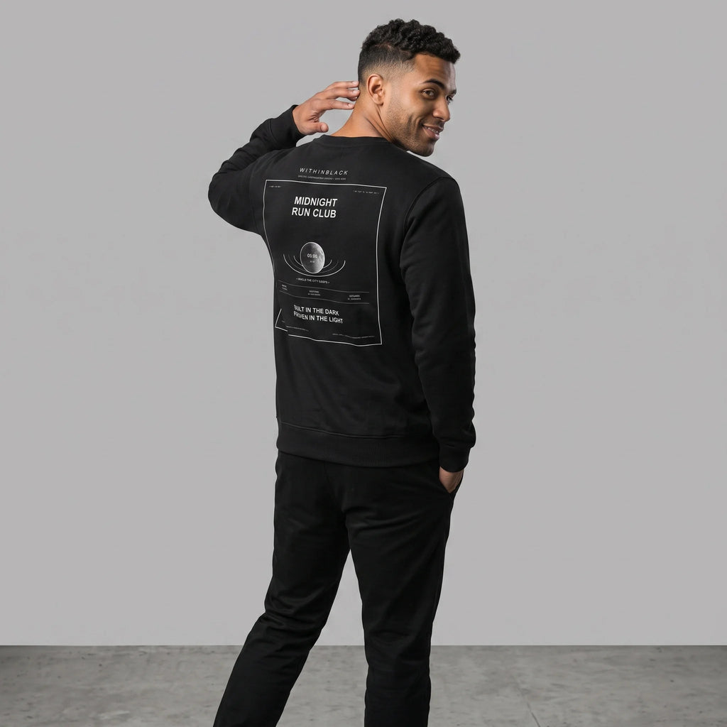 Three-quarter back view of a smiling male model turning toward the camera with one hand near his neck, wearing an all-black outfit including a black crewneck sweatshirt with the 'MIDNIGHT RUN CLUB' back graphic print.