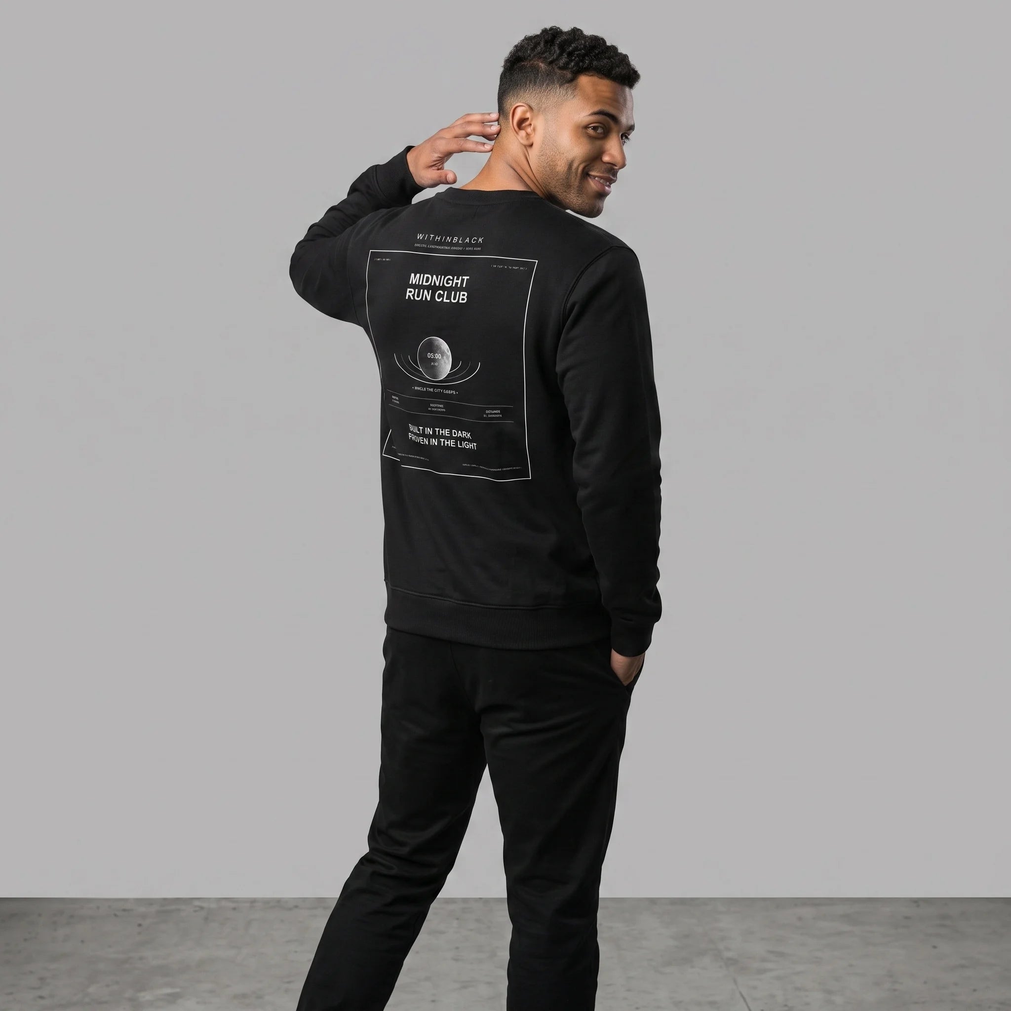 Three-quarter back view of a smiling male model turning toward the camera with one hand near his neck, wearing an all-black outfit including a black crewneck sweatshirt with the 'MIDNIGHT RUN CLUB' back graphic print.