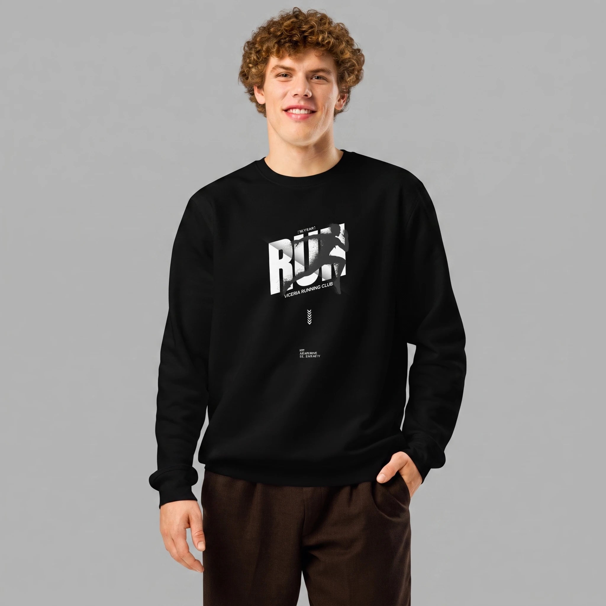 A smiling male model with curly brown hair standing against a grey background with one hand in his pocket. He is wearing a black crewneck sweatshirt paired with dark brown trousers. The sweatshirt features a white and grey graphic on the chest with the bold text 'RUN', a runner's silhouette across the letters, and 'VIENNA RUNNING CLUB' printed below.