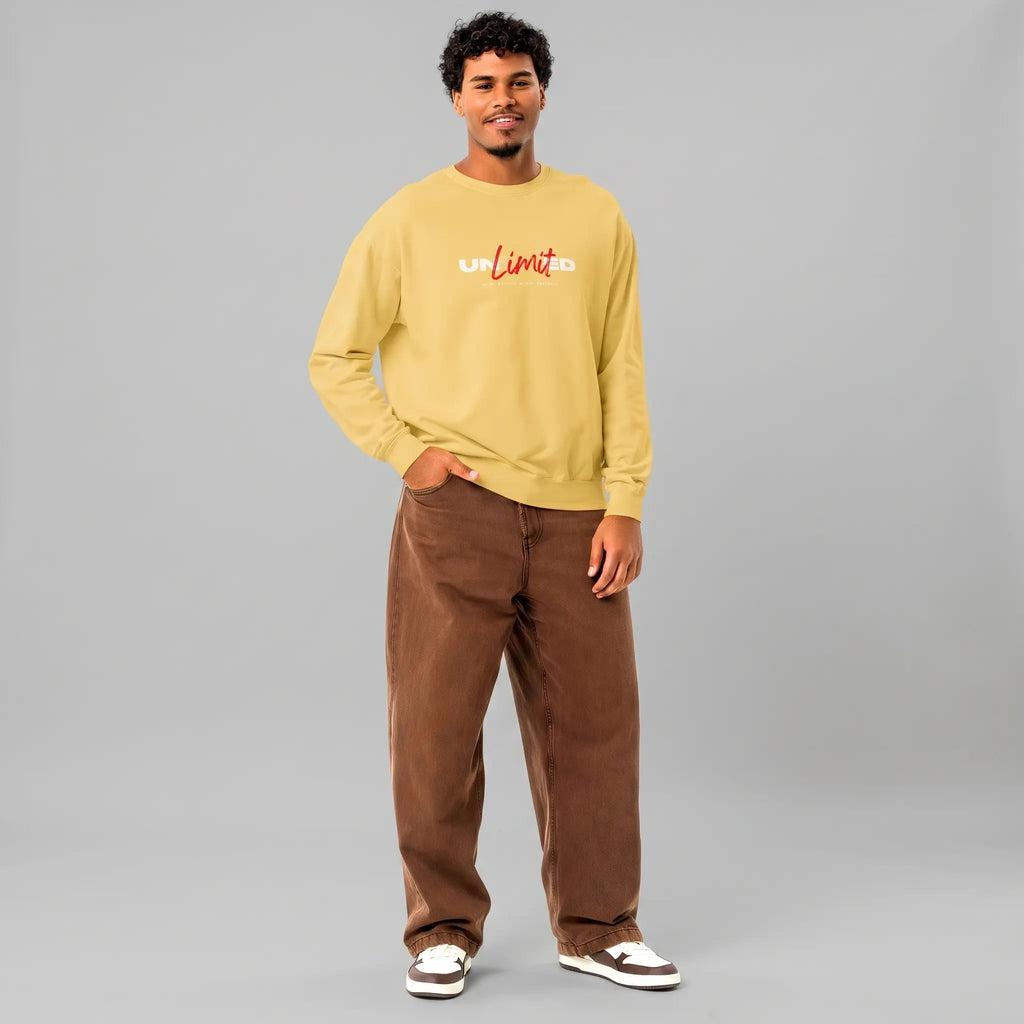 Full-length front view of a smiling male model with short curly hair and a goatee, standing against a grey background with one hand in his pocket. He is wearing a yellow crewneck sweatshirt with 'UNLIMITED' printed on the chest in white block letters with 'Limit' in red script, styled with baggy brown jeans and white sneakers with brown details.