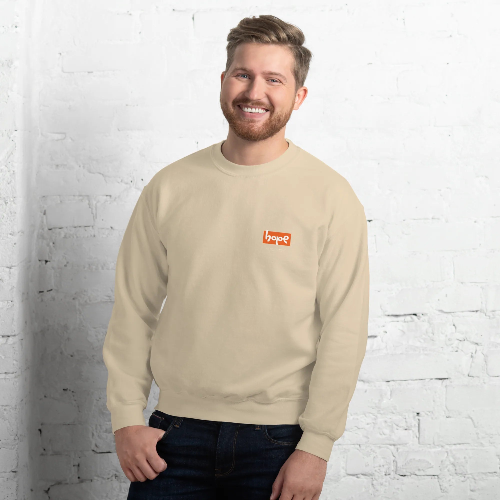 Smiling male model standing against white brick wall wearing sand beige crewneck sweatshirt with orange Hope chest logo front view