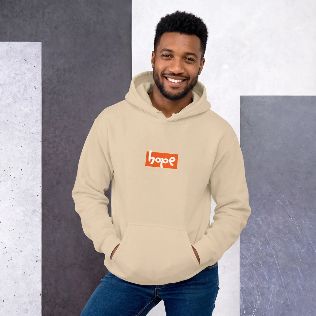 Smiling Black model wearing a sand-colored pullover hoodie with an orange rectangular 'hope' box logo, against a geometric wall.