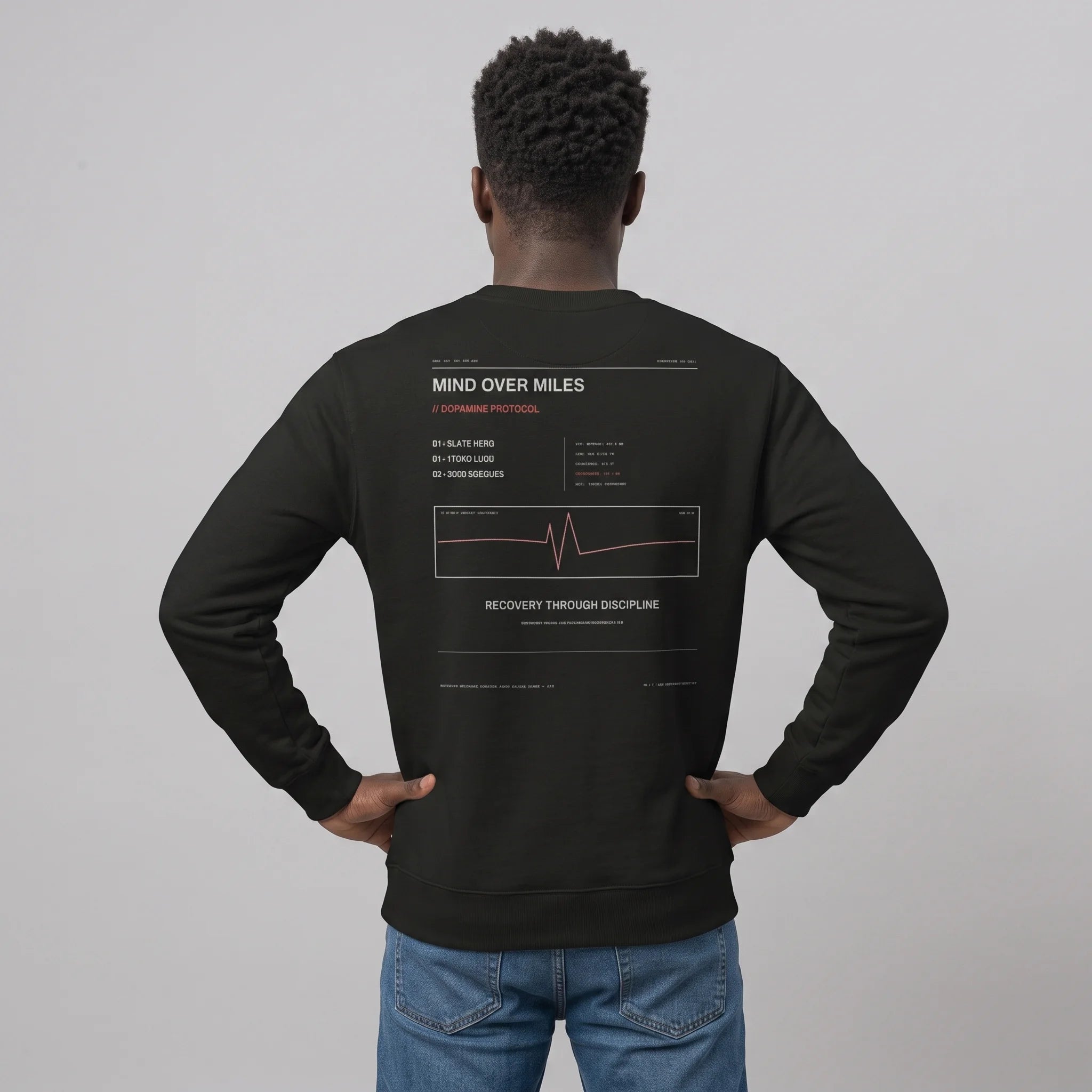 Back view of a male model standing with hands on hips against a grey studio background, wearing a black crewneck sweatshirt featuring the 'MIND OVER MILES / DOPAMINE PROTOCOL' graphic print with an EKG line, paired with blue jeans.