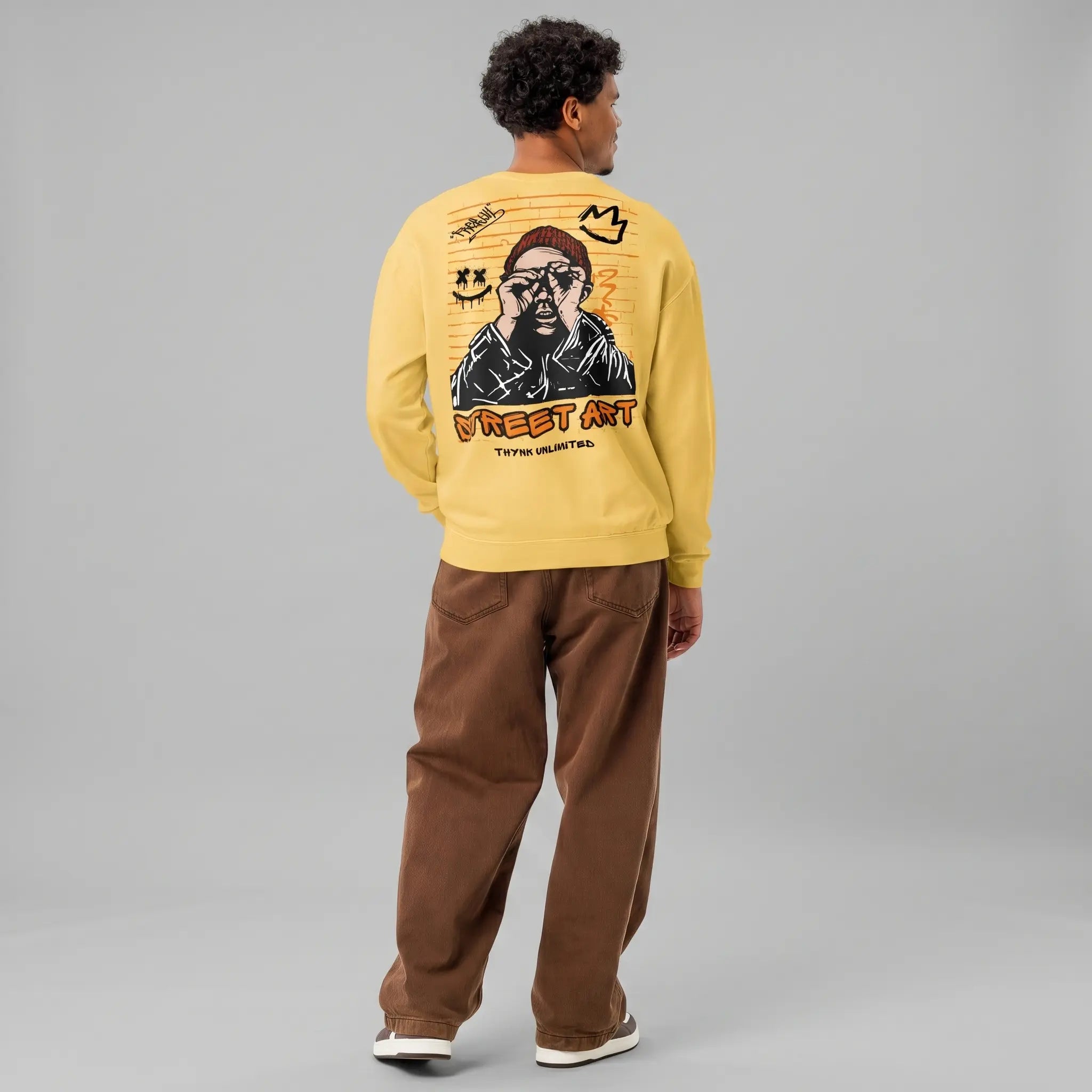 A back view of a male model with dark curly hair, wearing a loose-fitting yellow crewneck sweatshirt and brown baggy jeans, standing against a grey background. The back of the sweatshirt features a detailed graffiti-style art graphic: a central drawing of a man in a red beanie peering through binoculars, surrounded by other small graffiti elements like a crown, a smiley face, and hand-drawn letters. Large, stylized orange and red letters below read 'STREET ART', and smaller text reads 'THYNK UNLIMITED'.