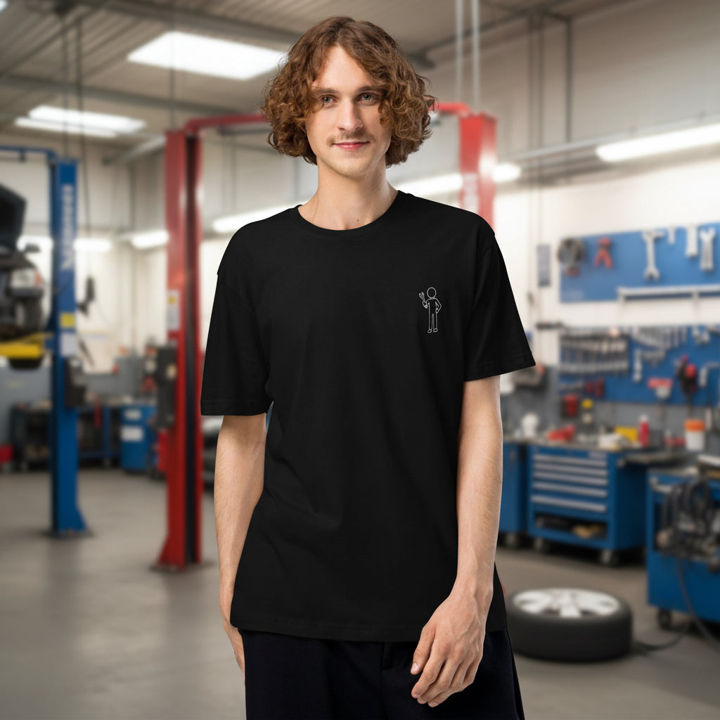 Male model standing in an auto repair garage wearing a black men's car enthusiast t-shirt with a small white mechanic graphic on the chest.