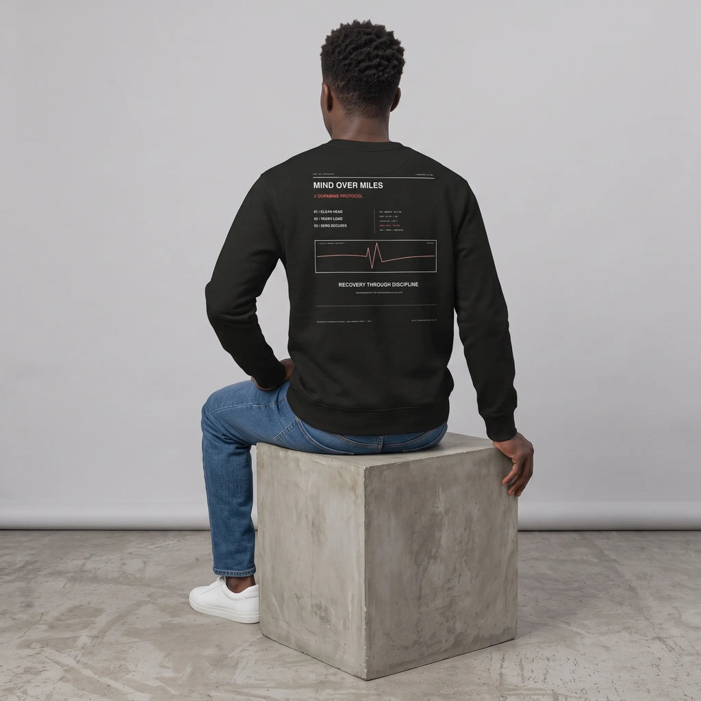 Back view of a male model seated on a concrete cube against a grey background, wearing a black crewneck sweatshirt with the 'MIND OVER MILES' and heartbeat line back print, paired with blue jeans and white sneakers.