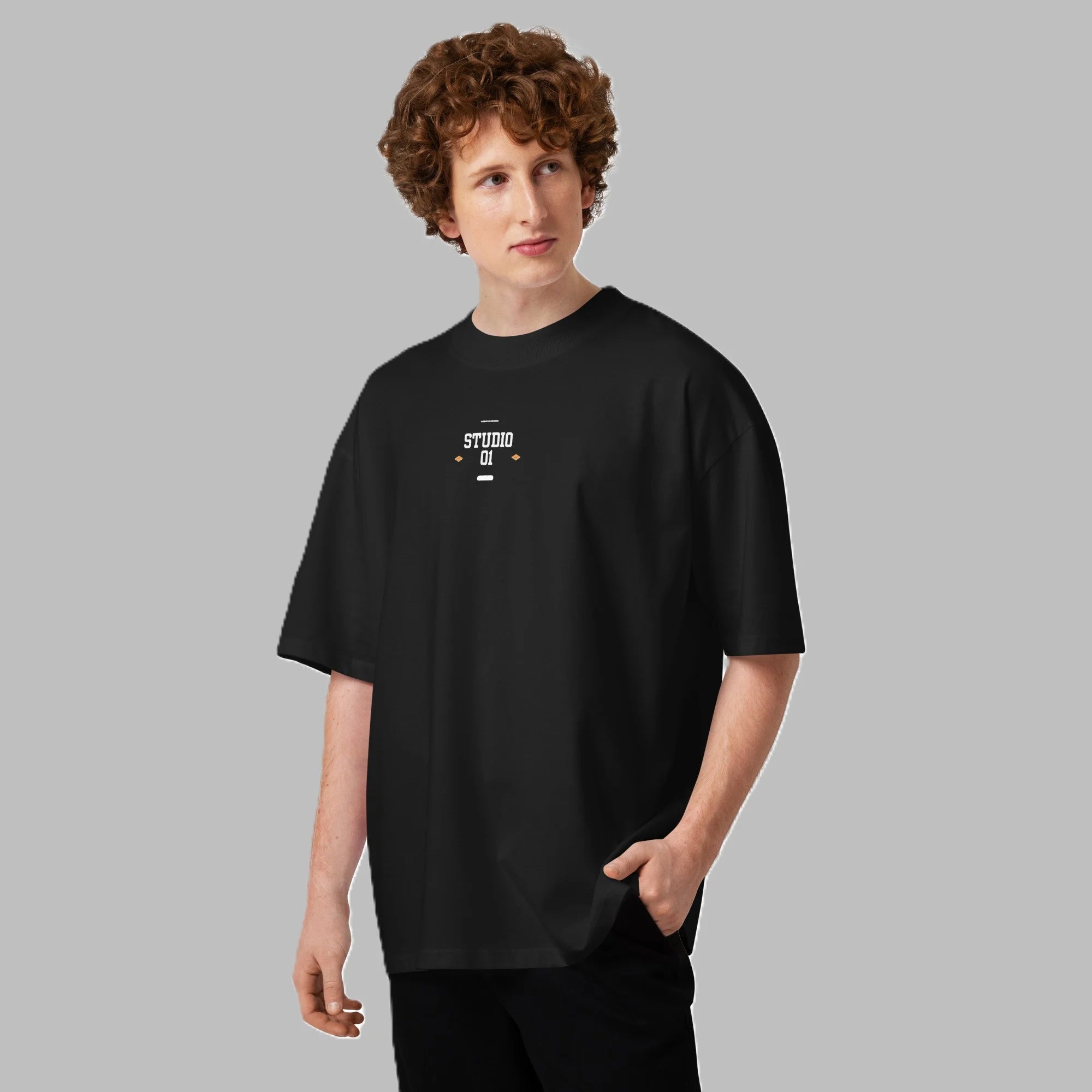 A male model from the front, standing with a hand in his pocket, wearing a black oversized heavyweight organic cotton streetwear t-shirt featuring a centered 'STUDIO 01' chest graphic.