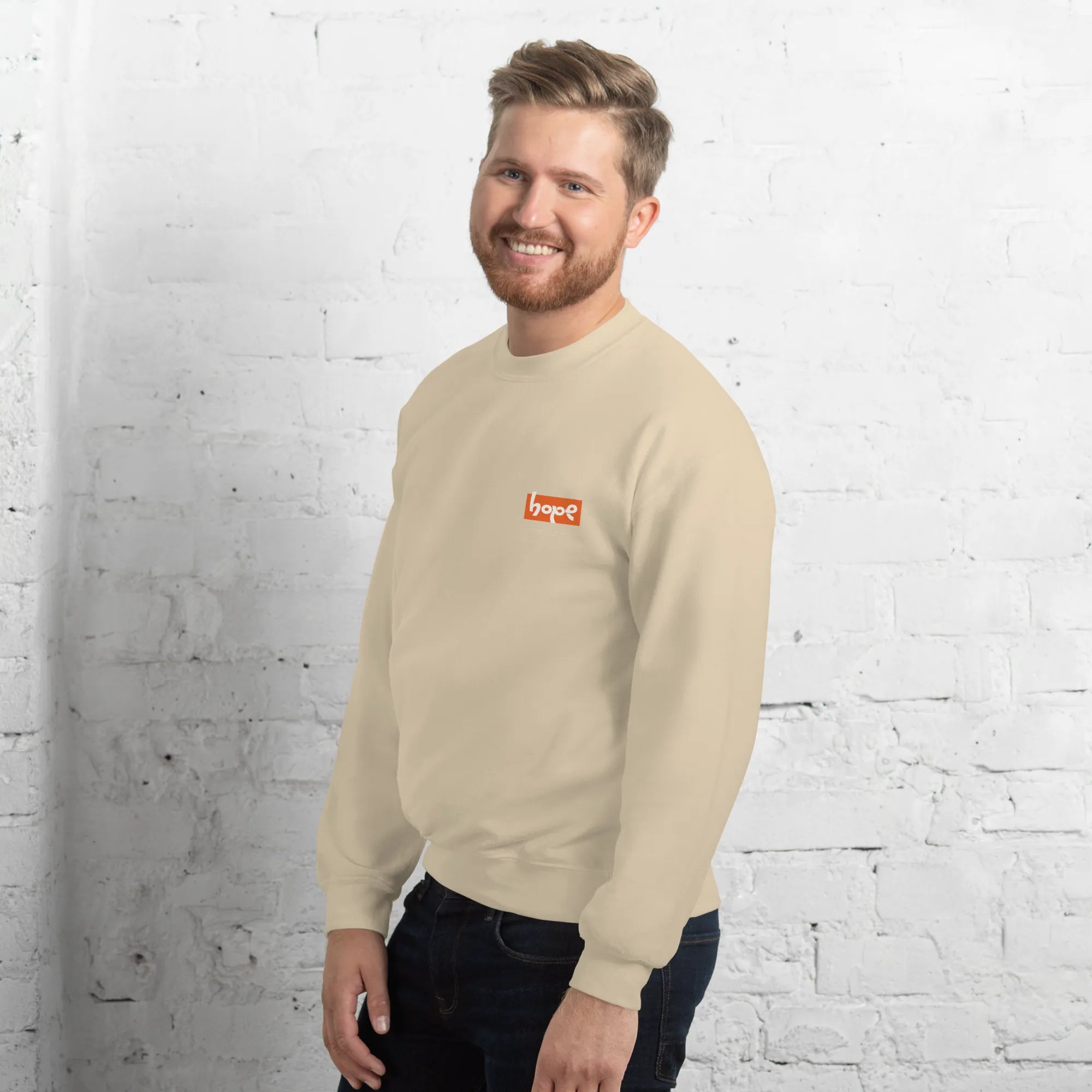 Side-angle view of smiling male model wearing sand beige crewneck sweatshirt with orange Hope chest logo against white brick wall