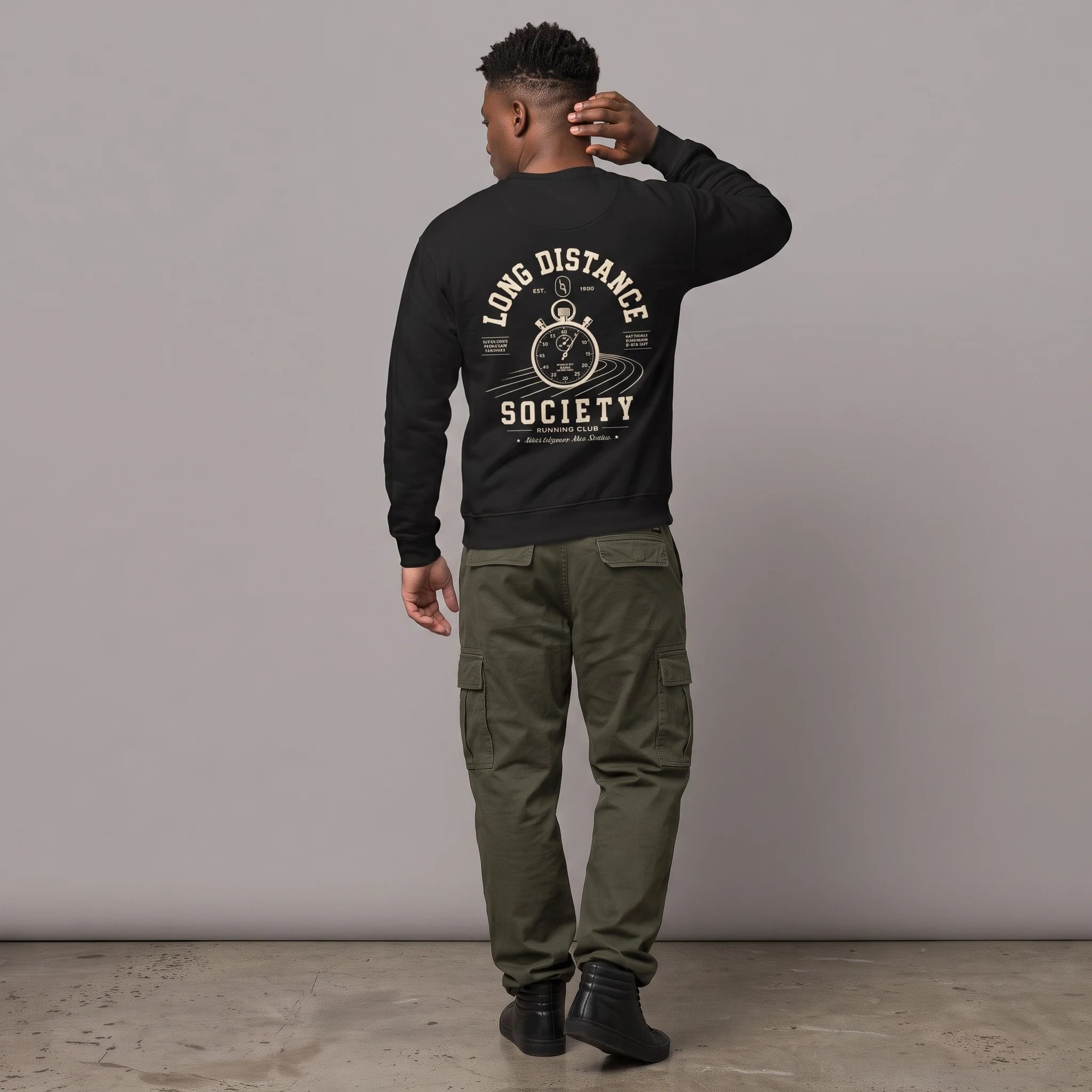 Full-length back view of a male model with short dark hair standing against a grey studio background. He is wearing a black crewneck sweatshirt paired with olive green cargo pants and black high-top sneakers. The back of the sweatshirt features an off-white graphic of a vintage stopwatch over running track lanes, surrounded by the text 'LONG DISTANCE SOCIETY RUNNING CLUB'.