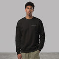 Front view of a male model with curly dark hair standing against a grey studio background with one hand in his pocket, wearing a black crewneck sweatshirt with 'WITHINBLACK MIDNIGHT ATHLETICS' printed on the chest, paired with olive green trousers.