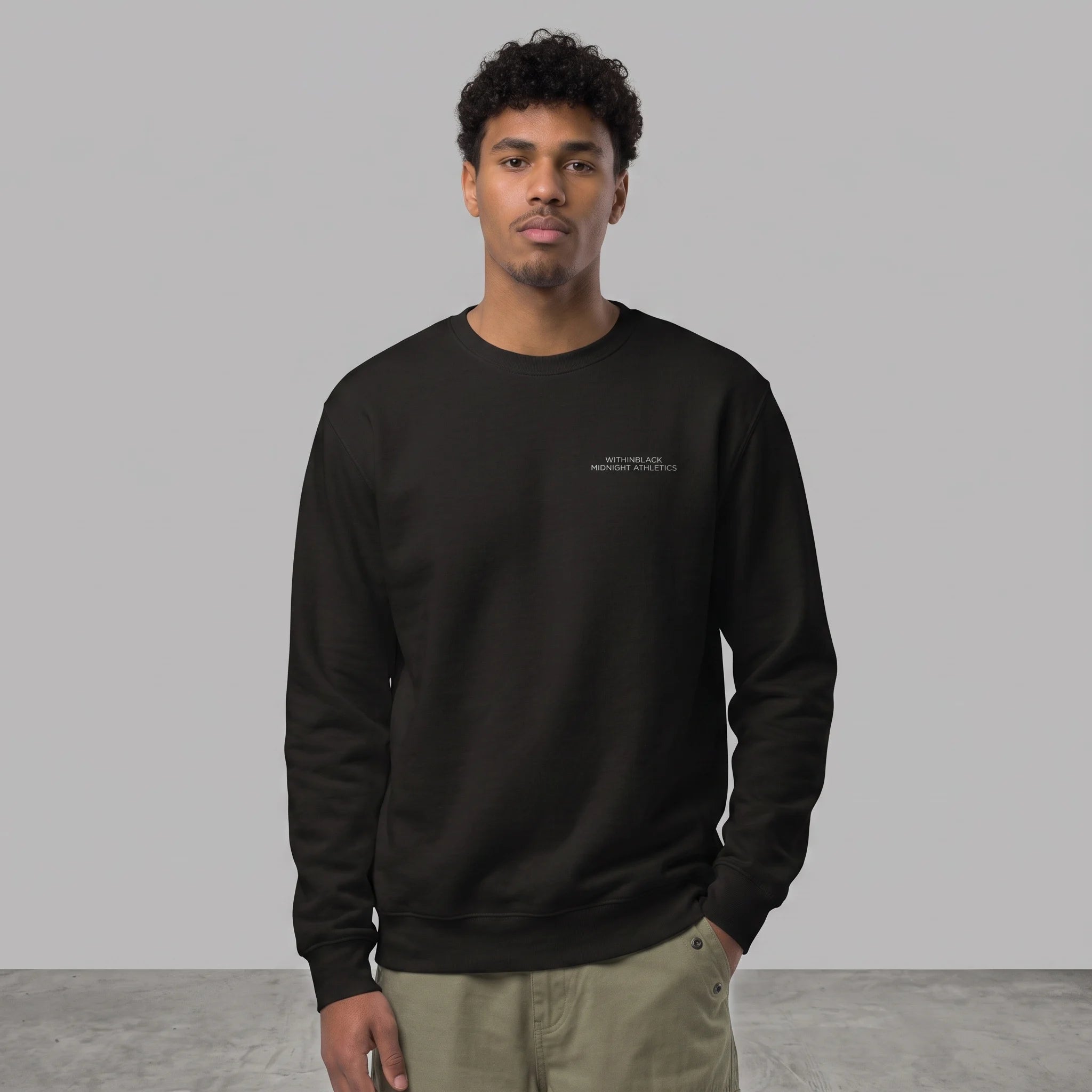 Front view of a male model with curly dark hair standing against a grey studio background with one hand in his pocket, wearing a black crewneck sweatshirt with 'WITHINBLACK MIDNIGHT ATHLETICS' printed on the chest, paired with olive green trousers.