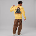 A male model seen from the back, wearing a yellow crewneck sweatshirt with a large street art graphic. The graphic includes a portrait of a man with hands on his face, graffiti elements, and the texts 'STREET ART' and 'THENK UNLIMITED'. He is also wearing loose brown pants.