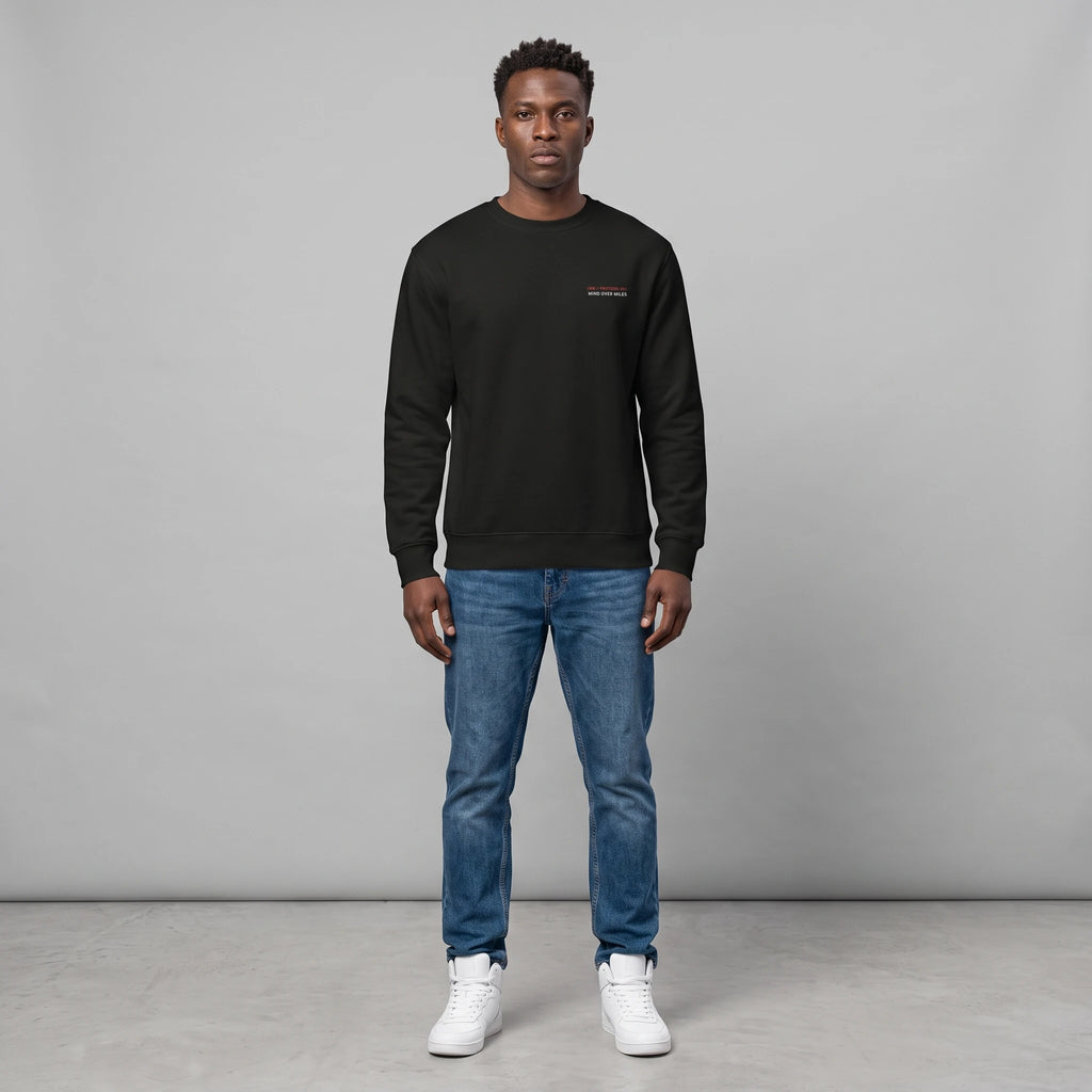 Full-length front view of a male model with short dark hair standing against a grey background in a studio setting, wearing a black crewneck sweatshirt with small chest text, paired with blue jeans and white sneakers.