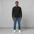 Full-length front view of a male model with short dark hair standing against a grey background in a studio setting, wearing a black crewneck sweatshirt with small chest text, paired with blue jeans and white sneakers.